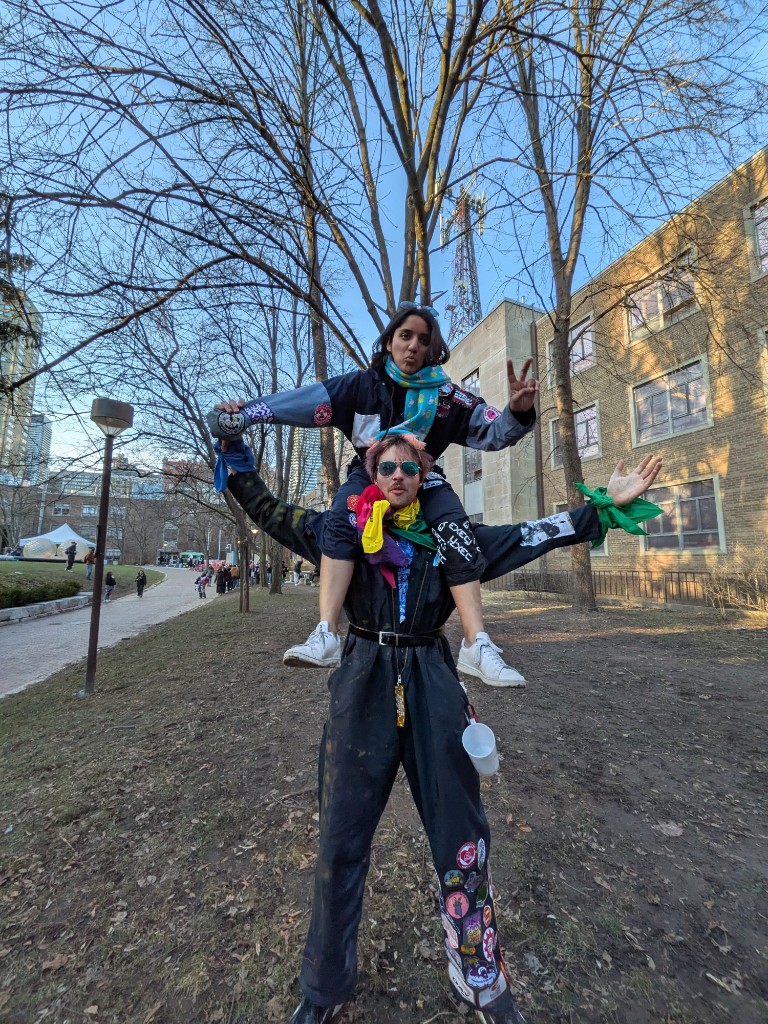 Two students in engineering coveralls, one sitting on the other's shoulders